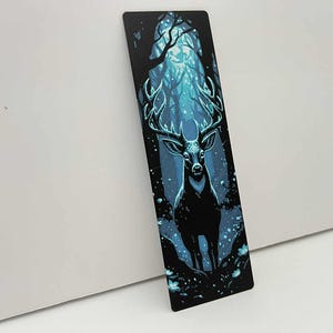 May include: A black bookmark featuring a majestic stag in a moonlit forest. The stag has large antlers and stands in a clearing with glowing blue light. The background shows a dense forest with a mystical, ethereal quality.