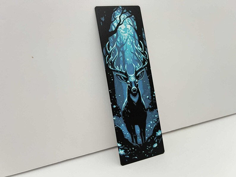 May include: A black bookmark featuring a majestic stag in a moonlit forest. The stag has large antlers and stands in a clearing with glowing blue light. The background shows a dense forest with a mystical, ethereal quality.
