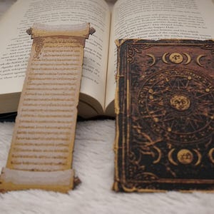 May include: An open book with a scroll-shaped bookmark and a decorative journal. The bookmark is tan with script and a rolled top and bottom. The journal is brown with a celestial design.
