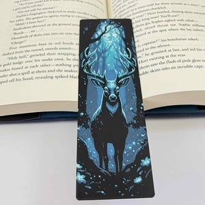 May include: A black bookmark featuring a majestic deer with large antlers, set against a blue and black forest scene. The bookmark is rectangular and is placed in an open book.