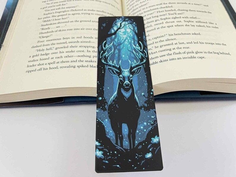 May include: A black bookmark featuring a majestic deer with large antlers, set against a blue and black forest scene. The bookmark is rectangular and is placed in an open book.
