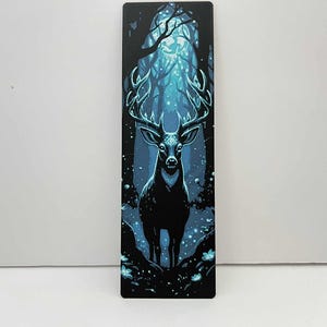 May include: A rectangular bookmark featuring a majestic stag in a mystical forest scene. The stag is black with large antlers, set against a backdrop of glowing blue trees and sparkling lights. The overall theme is fantasy and nature.