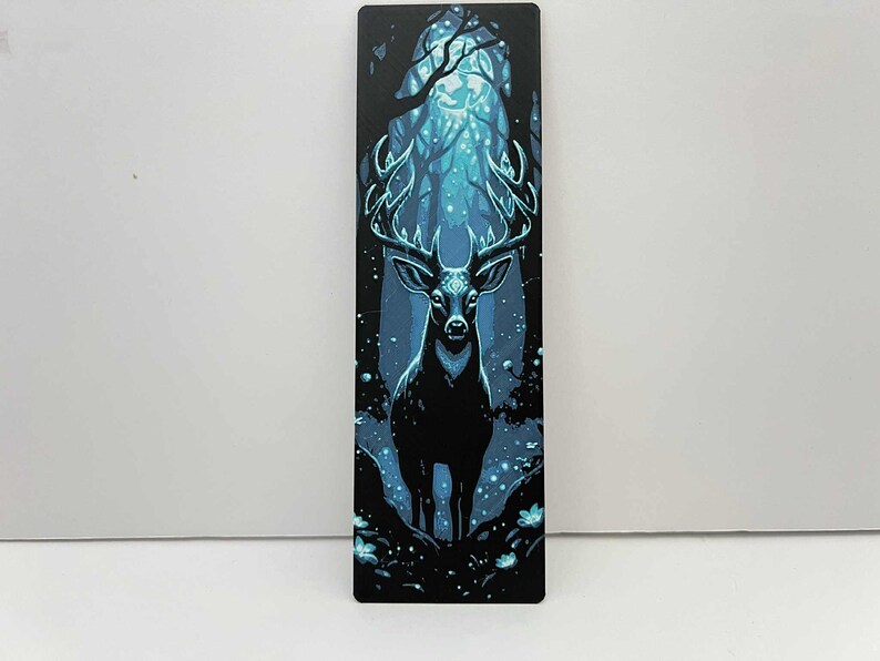 May include: A rectangular bookmark featuring a majestic stag in a mystical forest scene. The stag is black with large antlers, set against a backdrop of glowing blue trees and sparkling lights. The overall theme is fantasy and nature.