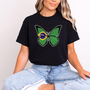 Puede incluir: Camiseta negra con un dise&ntilde;o de mariposa. Las alas de la mariposa son verdes con contornos negros, y el centro de las alas muestra los colores de la bandera brasile&ntilde;a: amarillo, azul y verde. La camiseta es de estilo casual.