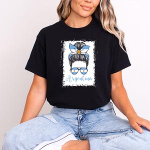 May include: Black t-shirt featuring a graphic of a woman with a blue and white Argentina flag-themed headband and sunglasses. The word "Argentina" is written in blue script below the image. The design is framed in a white rectangle.