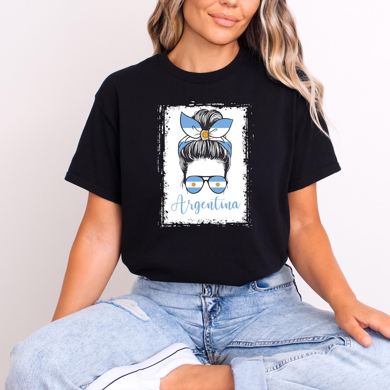 May include: Black t-shirt featuring a graphic of a woman with a blue and white Argentina flag-themed headband and sunglasses. The word "Argentina" is written in blue script below the image. The design is framed in a white rectangle.