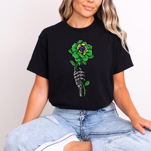 Puede incluir: Camiseta negra con una rosa verde y amarilla sostenida por una mano esquel&eacute;tica. La rosa tiene p&eacute;talos verdes y un centro amarillo y azul. El dise&ntilde;o es una ilustraci&oacute;n gr&aacute;fica.