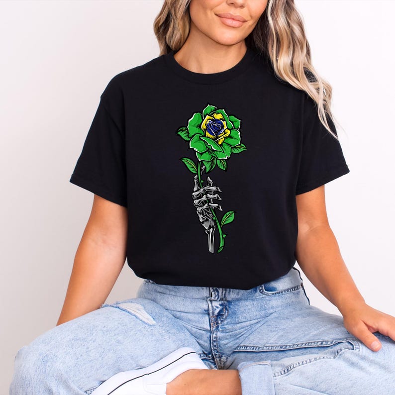 Puede incluir: Camiseta negra con una rosa verde y amarilla sostenida por una mano esquel&eacute;tica. La rosa tiene p&eacute;talos verdes y un centro amarillo y azul. El dise&ntilde;o es una ilustraci&oacute;n gr&aacute;fica.