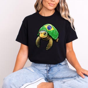 Puede incluir: Camiseta negra con una ilustraci&oacute;n detallada de una tortuga. La tortuga tiene un caparaz&oacute;n verde con el dise&ntilde;o de la bandera brasile&ntilde;a, una cara amarilla y patas marrones. La camiseta est&aacute; hecha de un material suave.