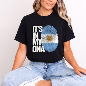 May include: Black t-shirt with the text "IT'S IN MY DNA" in white. The design includes a fingerprint in the colors of the Argentinian flag. The shirt is a short-sleeved crew neck.