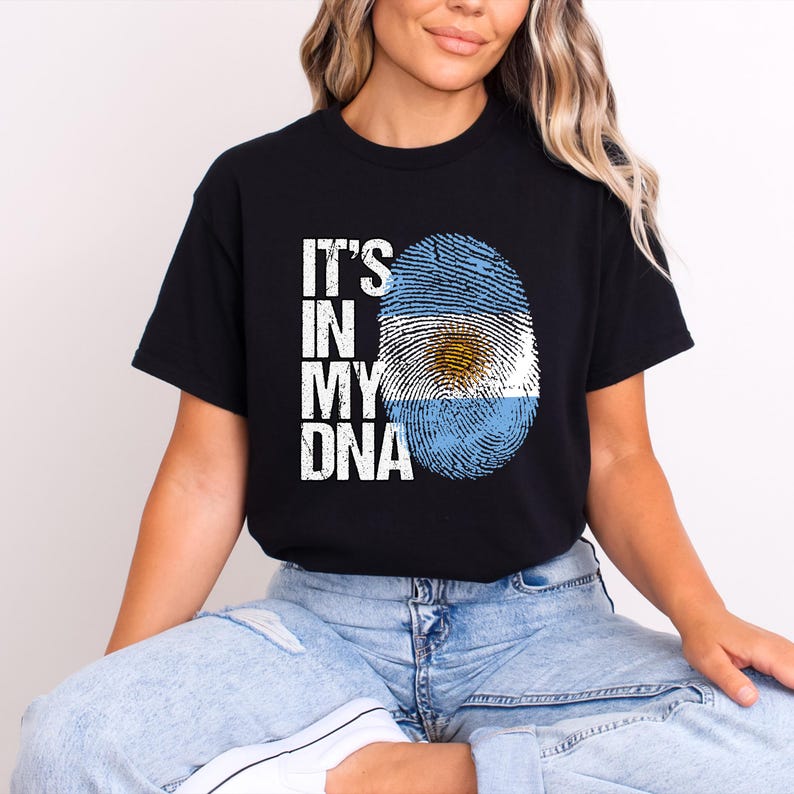 May include: Black t-shirt with the text "IT'S IN MY DNA" in white. The design includes a fingerprint in the colors of the Argentinian flag. The shirt is a short-sleeved crew neck.