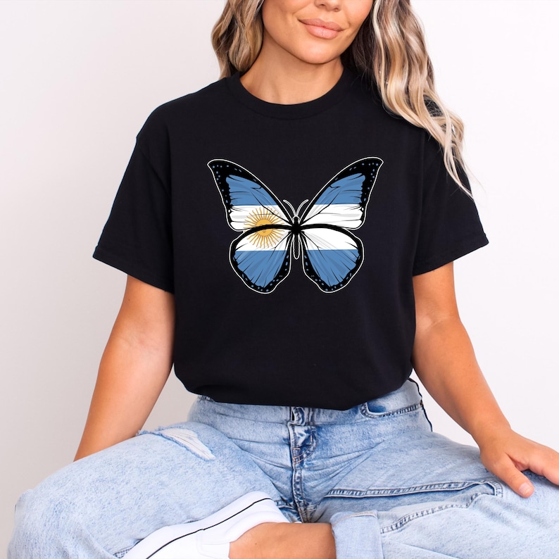 May include: Black t-shirt featuring a butterfly design with the Argentinian flag. The butterfly has blue and white wings with a yellow sun in the center. The shirt is a classic crew neck style.