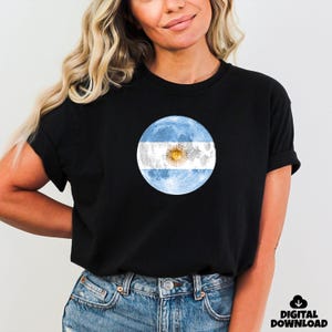 May include: Black t-shirt featuring a circular design of the Argentinian flag, with light blue stripes and a sun in the center. The words "DIGITAL DOWNLOAD" are in the lower right corner.