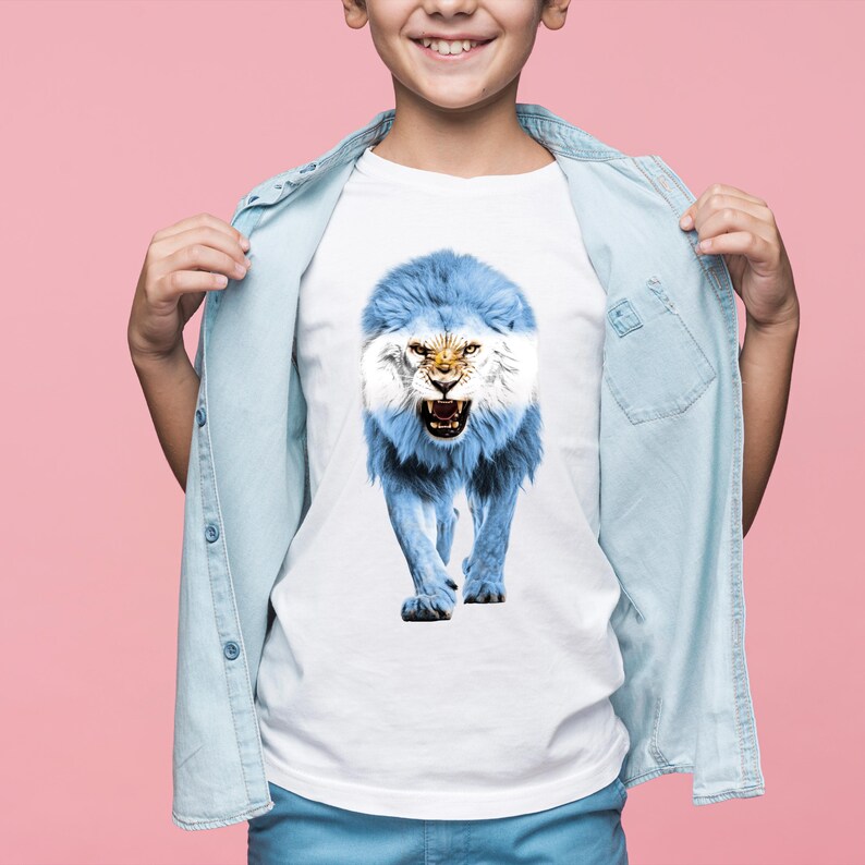 May include: White t-shirt featuring a roaring lion design in the colors of the Argentinian flag: light blue and white. The lion's mane and body are light blue, with the face and details in white. The shirt is worn by a person with a light blue denim shirt.
