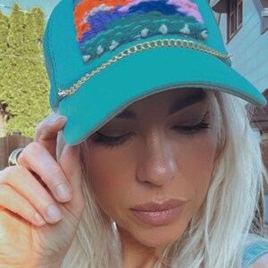 May include: Teal trucker hat with a woven patch featuring colorful abstract design. The hat has a gold chain detail along the brim. The hat is a stylish accessory.