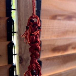 May include: A handmade macrame keychain in a deep red hue, featuring a braided design and accented with dark wooden beads. The keychain is suspended against a blurred background of wooden slats and black hinges.