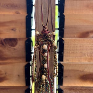 May include: Handmade macrame pendant necklace featuring a central woven design with tan, green, and brown threads. The necklace includes wooden beads and a brown leather cord, creating a bohemian aesthetic.