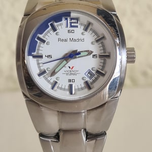 May include: A stainless steel wristwatch with a white face and blue accents. The watch features the text "Real Madrid" and a date display. The watch band is also stainless steel.