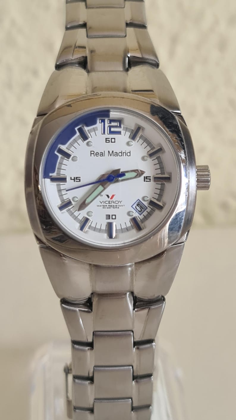 May include: A stainless steel wristwatch with a white face and blue accents. The watch features the text "Real Madrid" and a date display. The watch band is also stainless steel.