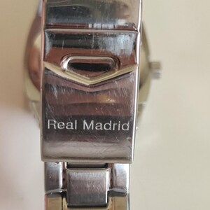 May include: Close-up of a silver-toned watch clasp with the words "Real Madrid" engraved on it. The clasp is rectangular with a hinged design, showing signs of wear. The watch band is partially visible.