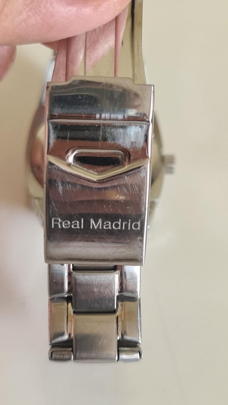 May include: Close-up of a silver-toned watch clasp with the words "Real Madrid" engraved on it. The clasp is rectangular with a hinged design, showing signs of wear. The watch band is partially visible.