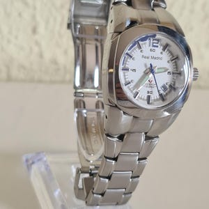 May include: A silver-toned wristwatch with a white face and a stainless steel band. The watch face displays the text "Real Madrid" and features a date window. The watch is displayed on a clear acrylic stand.