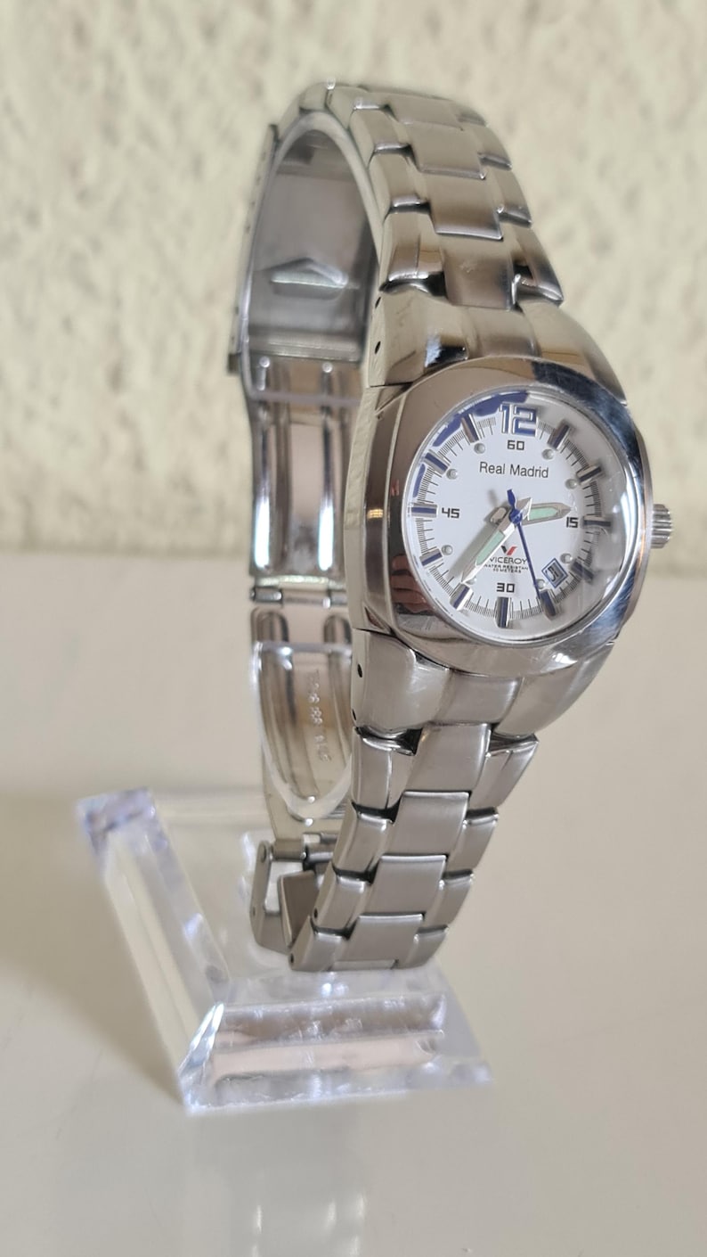 May include: A silver-toned wristwatch with a white face and a stainless steel band. The watch face displays the text "Real Madrid" and features a date window. The watch is displayed on a clear acrylic stand.