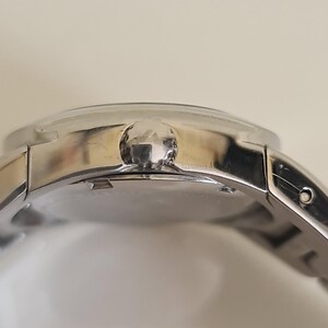 May include: Close-up of a silver wristwatch with a round face and a silver band. The watch has a textured crown and a polished finish. The watch is a classic accessory.