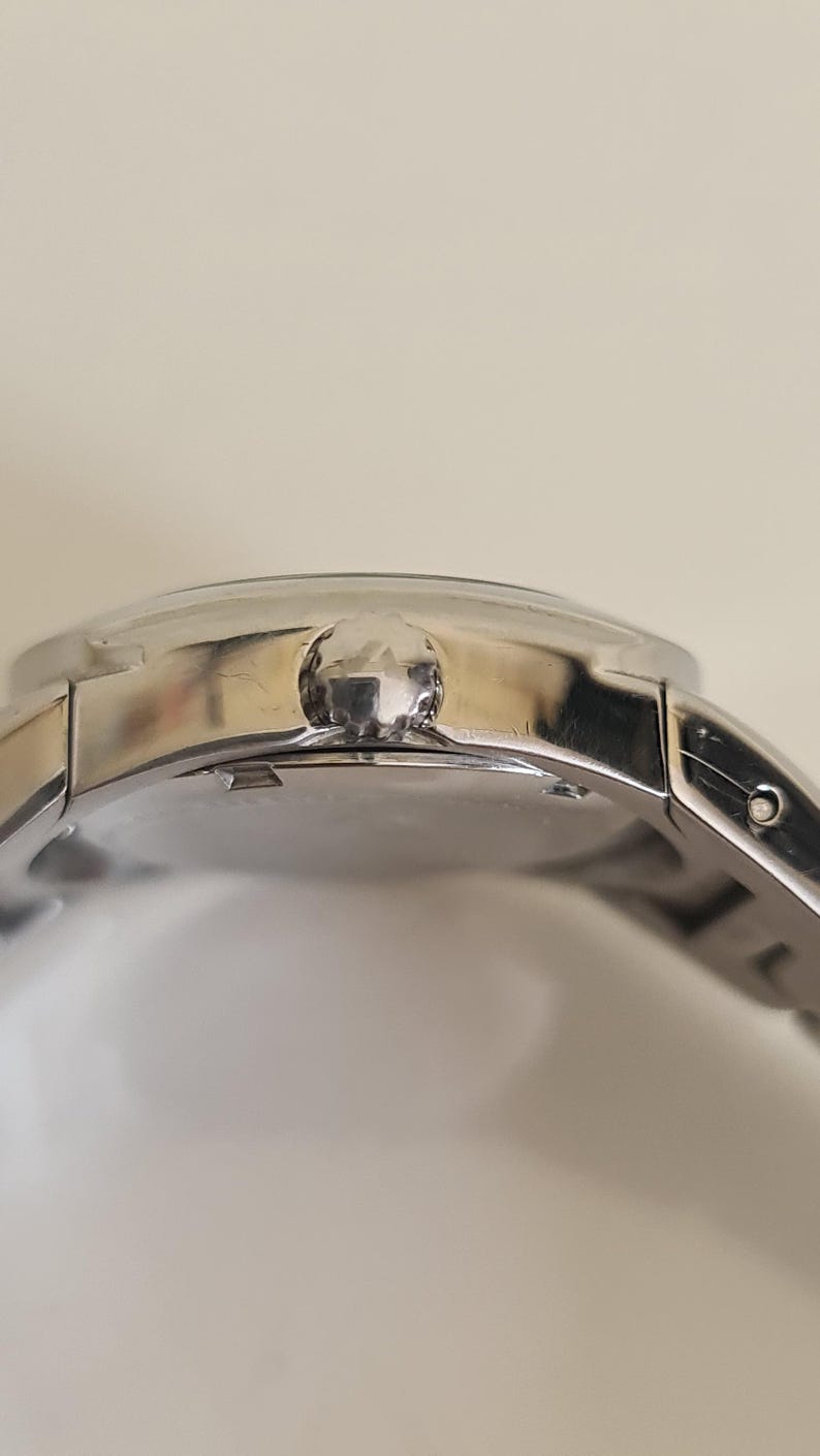 May include: Close-up of a silver wristwatch with a round face and a silver band. The watch has a textured crown and a polished finish. The watch is a classic accessory.