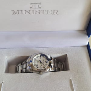 May include: A silver-toned Minister watch with a white face and Roman numeral hour markers, presented in a blue and white box. The watch has a stainless steel band and is new in its original packaging.