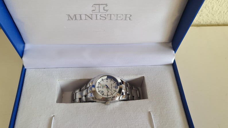 May include: A silver-toned Minister watch with a white face and Roman numeral hour markers, presented in a blue and white box. The watch has a stainless steel band and is new in its original packaging.
