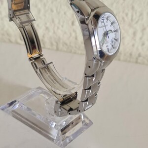 May include: A silver-toned wristwatch with a white face and a stainless steel band. The watch is displayed on a clear stand, showcasing its design. The watch face has black numerals and hands. The band is made of small, connected metal links.