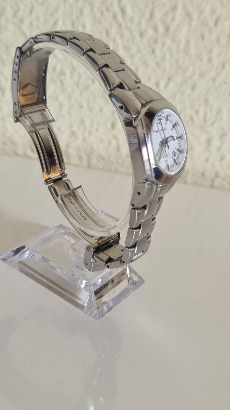 May include: A silver-toned wristwatch with a white face and a stainless steel band. The watch is displayed on a clear stand, showcasing its design. The watch face has black numerals and hands. The band is made of small, connected metal links.