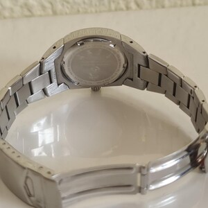 May include: A silver-toned wristwatch with a metal band, viewed from the back. The watch band is open, revealing the clasp. The watch face is visible, with a circular design and text. The watch is on a white surface.