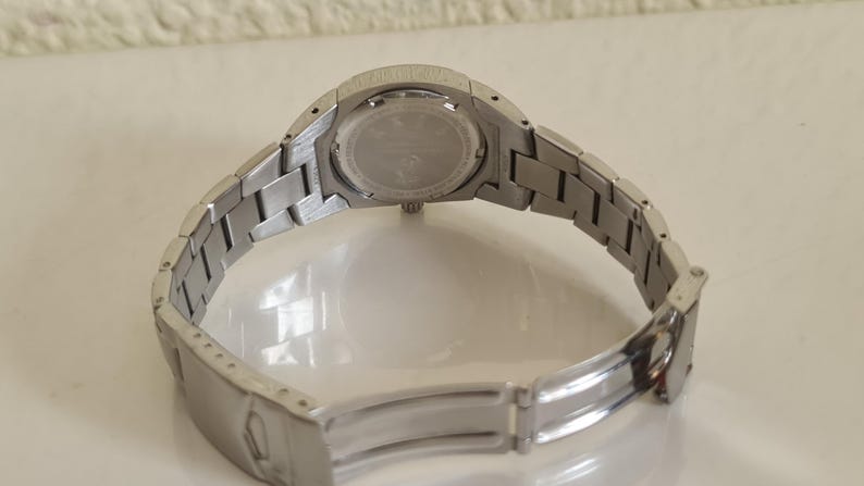 May include: A silver-toned wristwatch with a metal band, viewed from the back. The watch band is open, revealing the clasp. The watch face is visible, with a circular design and text. The watch is on a white surface.