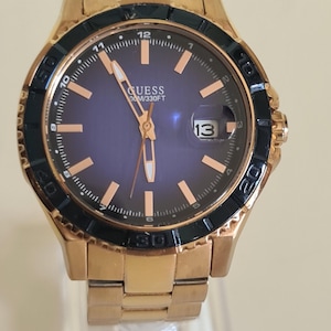 May include: A gold-coloured Guess watch with a dark blue dial and black bezel. The watch features gold hands and markers, a date display at the 3 o'clock position, and a gold link bracelet. The watch is marked "GUESS 100M/330FT".