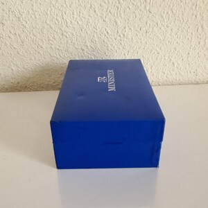 May include: A rectangular, cobalt blue box with the word "MINISTER" printed in white. The box is sitting on a white surface.