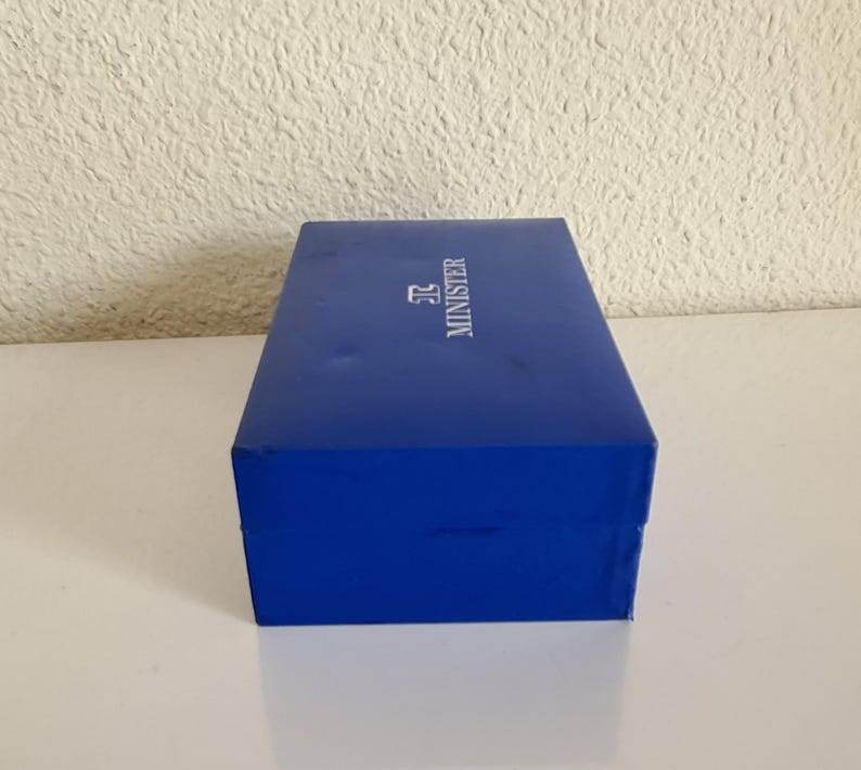 May include: A rectangular, cobalt blue box with the word "MINISTER" printed in white. The box is sitting on a white surface.