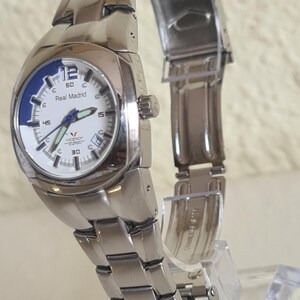 May include: A silver-toned wristwatch with a white face and blue accents. The watch features the text "Real Madrid" and the brand "Viceroy". The watch has a stainless steel band and is displayed on a clear stand.