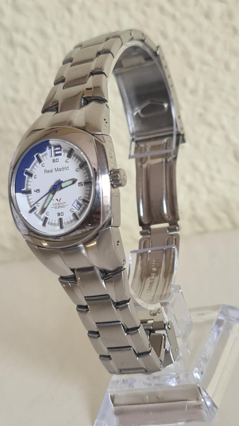 May include: A silver-toned wristwatch with a white face and blue accents. The watch features the text "Real Madrid" and the brand "Viceroy". The watch has a stainless steel band and is displayed on a clear stand.