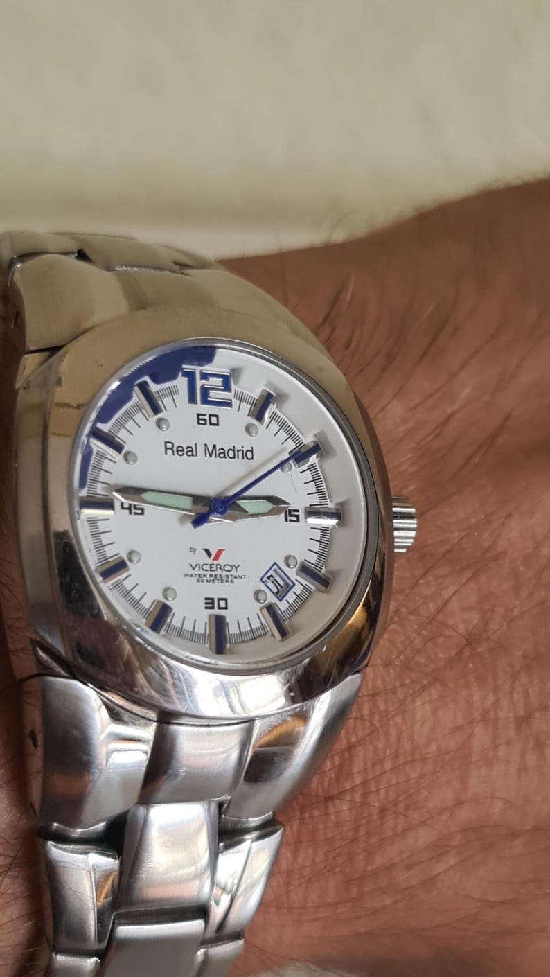 May include: Silver-toned Viceroy watch with a white face and blue accents. The watch face displays the text "Real Madrid" and "Viceroy Water Resistant 50 Meters". The watch has a date display at the 3 o'clock position and a silver-toned band.