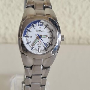 May include: A silver-toned wristwatch with a white face and the words "Real Madrid" printed on the dial. The watch has a stainless steel band and a blue bezel. The watch is displayed on a clear stand.