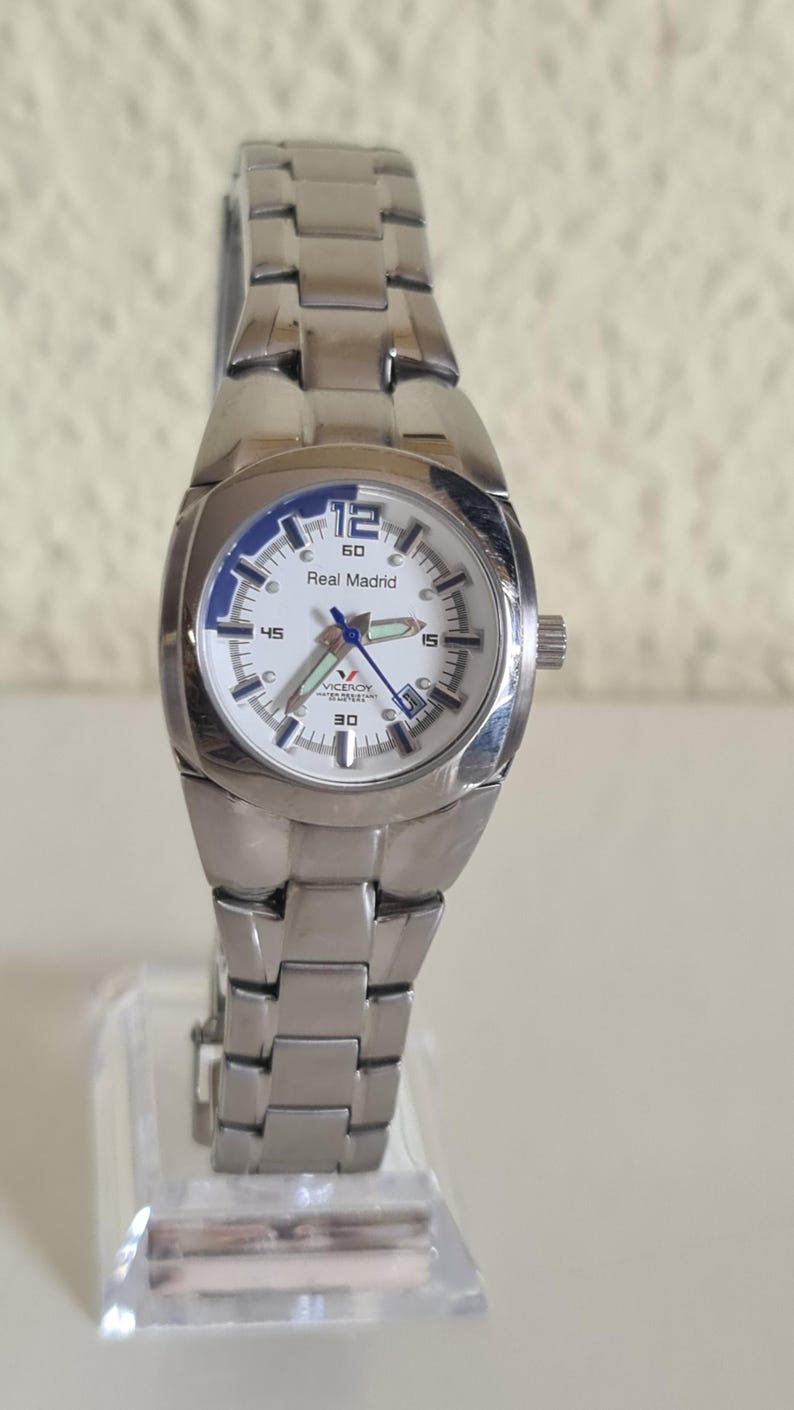 May include: A silver-toned wristwatch with a white face and the words "Real Madrid" printed on the dial. The watch has a stainless steel band and a blue bezel. The watch is displayed on a clear stand.