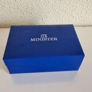 May include: A rectangular, cobalt blue box with the word "MINISTER" in white lettering on the lid. The box is closed and sits on a white surface, reflecting the box's color. The box appears to be made of a sturdy material.