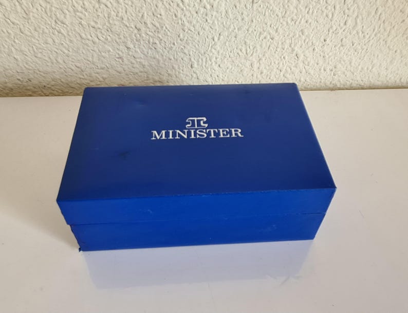 May include: A rectangular, cobalt blue box with the word "MINISTER" in white lettering on the lid. The box is closed and sits on a white surface, reflecting the box's color. The box appears to be made of a sturdy material.