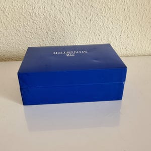 May include: A rectangular, cobalt blue box with the word "MINISTER" printed in silver on the lid. The box is closed and sits on a white surface, reflecting the box's shape. The background is a textured, off-white wall.