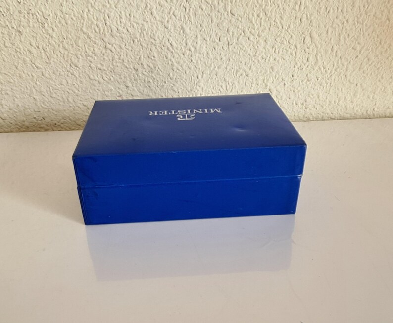 May include: A rectangular, cobalt blue box with the word "MINISTER" printed in silver on the lid. The box is closed and sits on a white surface, reflecting the box's shape. The background is a textured, off-white wall.