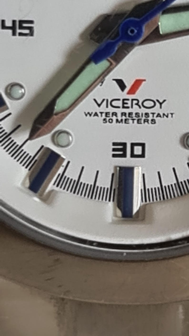 May include: Close-up of a white Viceroy watch face. The watch features black and blue hands, a black minute track, and the text "WATER RESISTANT 50 METERS". The number "30" is prominently displayed.