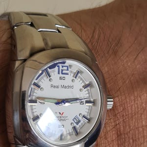 May include: A silver-toned wristwatch with a white face featuring the text "Real Madrid" and the number "12". The watch has a stainless steel band and a date display. The watch is a Viceroy brand.
