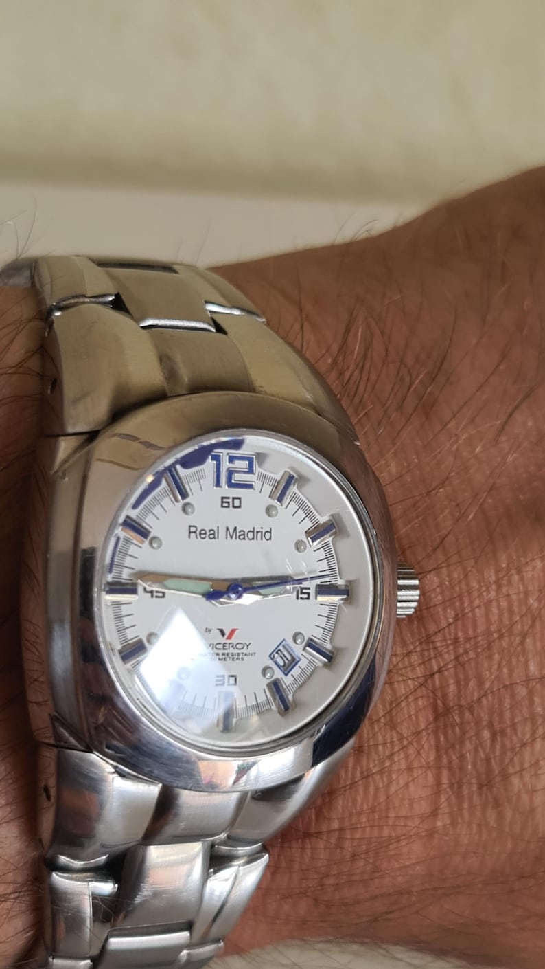 May include: A silver-toned wristwatch with a white face featuring the text "Real Madrid" and the number "12". The watch has a stainless steel band and a date display. The watch is a Viceroy brand.